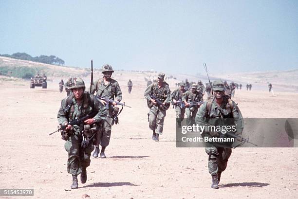 Grenada: United States combat soldiers run in parallel lines across an empty dirt field with their rifles at ready during the United States invasion...