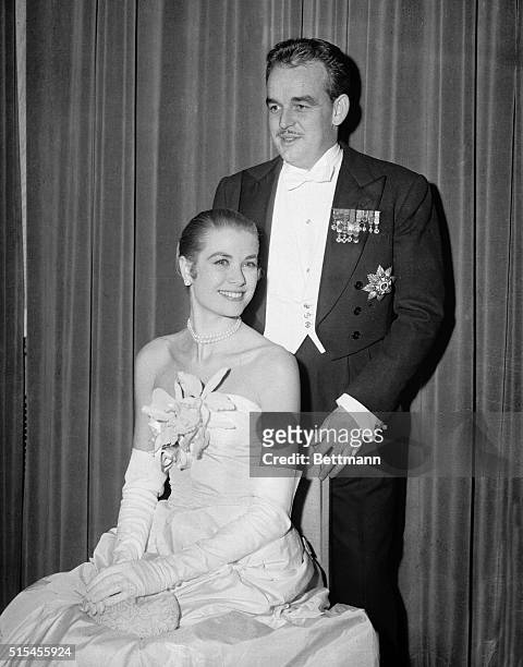 Bemedalled Prince Rainier III of Monaco stands behind the chair of his fiancee, actress Grace Kelly, attired in a decollet white, floor-length ball...