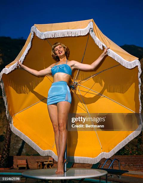 Actress Dorothy Abbott stands atop a table with a patio table umbrella while modeling a sky blue bikini in Hollywood.