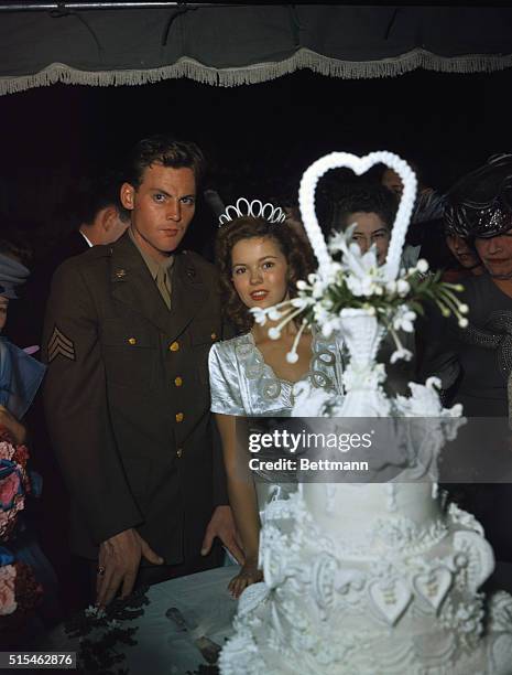 Shirley Temple is shown peeking out from behind the large wedding cake as her new husband, Sgt. Jack Agar, looks on with a serious expression.