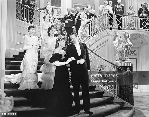 Greta Garbo as Anna Karenina walking down staircase on the arm of husband, Alexei Karenina played by Basil Rathbone. Behind them is a procession of...