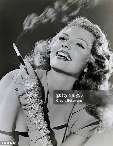 Publicity close up of Hollywood actress Rita Hayworth wearing ornately decorated gloves and holding a cigarette in a cigarette holder.