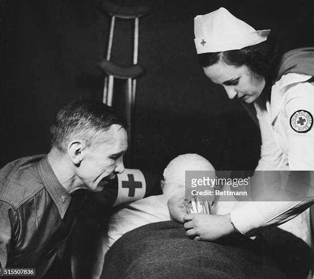 An American Red Cross nurse gives water to a wounded soldier head and eyes are bangaged as another man looks on; there are crutches in the background.