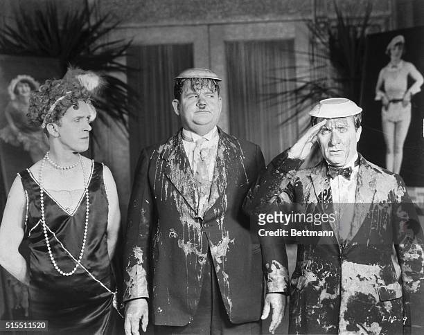 Stan Laurel, wearing a dress, looks at Oliver Hardy and another actor covered with pie in a movie still.