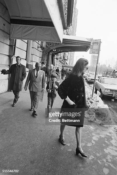 Jackie Onassis walks along a sidewalk by the Plaza Hotel, followed by her husband Aristotle and her son, JFK Jr.