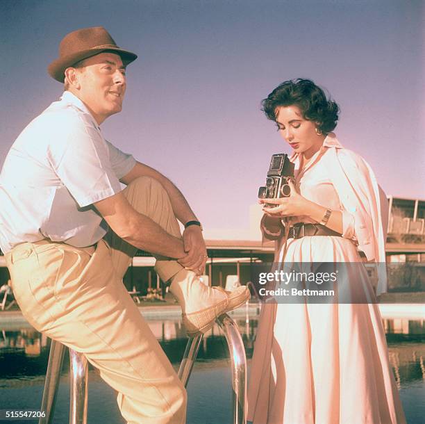 Actress Elizabeth Taylor is examining a Rolliflex camera while husband Michael Wilding relaxes at poolside.