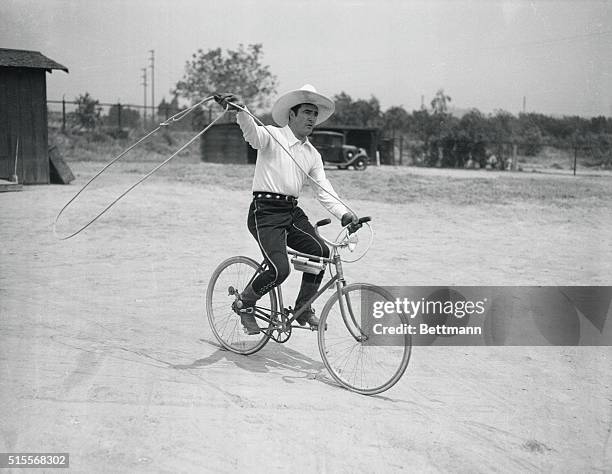 Famous cowboy star of the Silent Era, Tom Mix, rides a bicycle swinging a lasso.