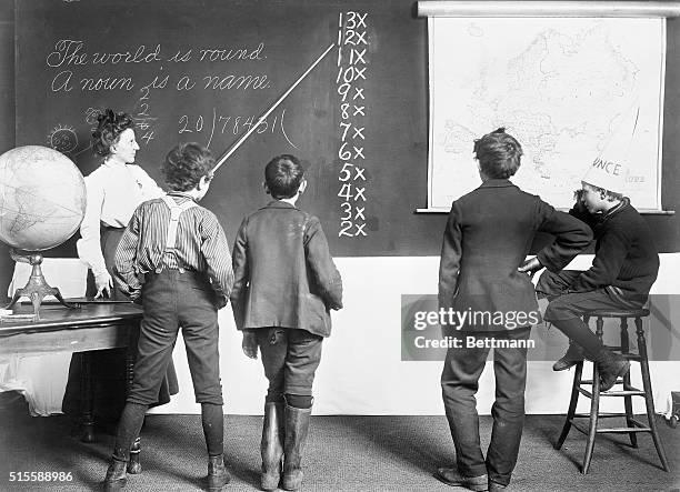 Four schoolboys watch as their teacher points to a lesson on the blackboard. The boy on the right is sitting wearing a dunce cap.