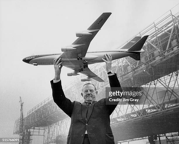 Keith Granville, Managing Director of BOAC, holding up a model of the Boeing 747 jet, with the new aircraft hangars under construction in the...