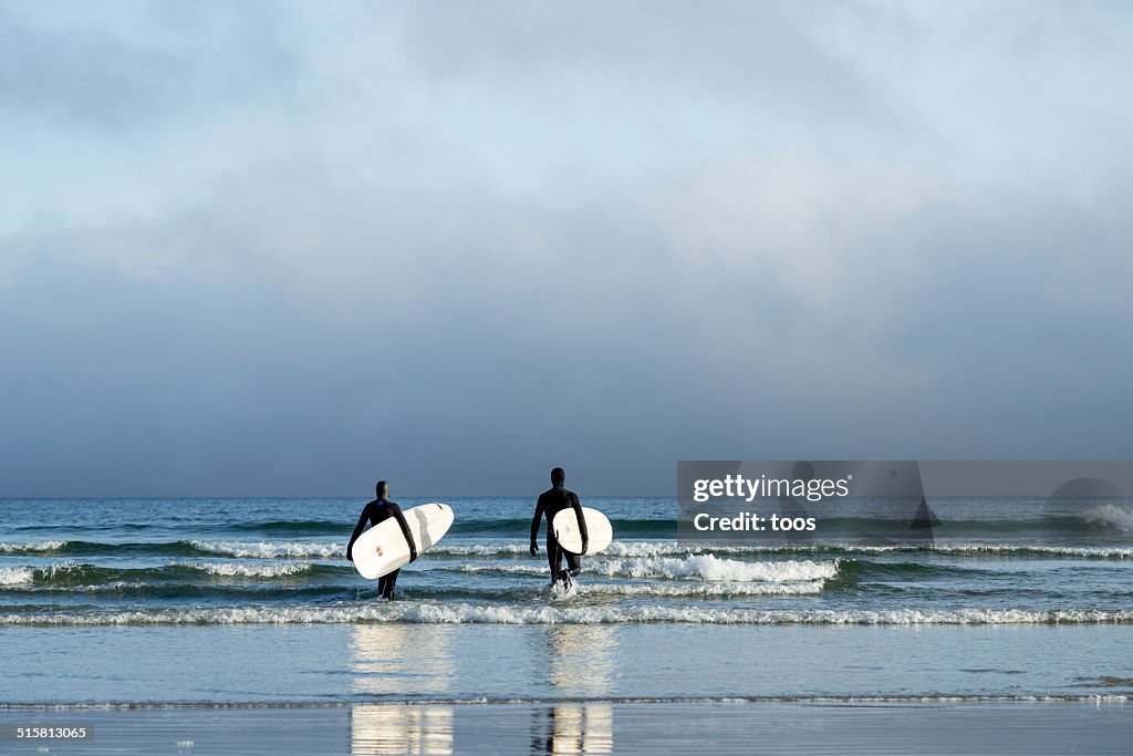 Two surfers in wetsuits walking into the Pacific Ocean