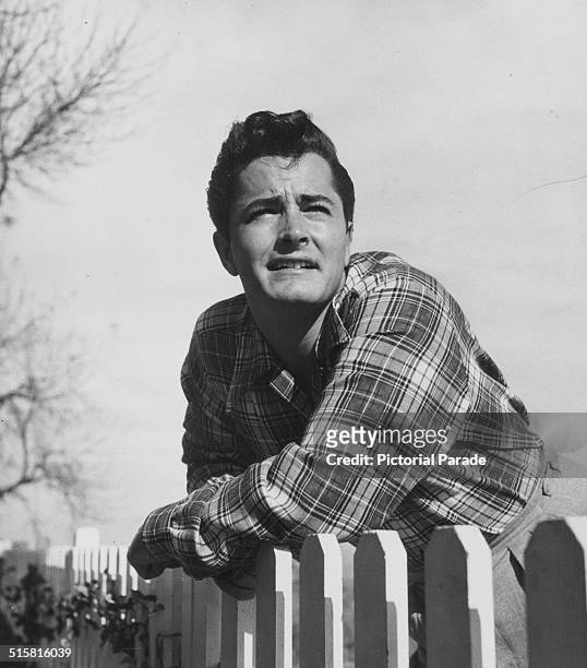 Portrait of actor John Derek, wearing a check shirt and leaning on a picket fence, circa 1950.