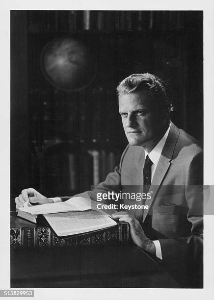 Portrait of American evangelist Billy Graham, sitting in a library with a book, circa 1970.