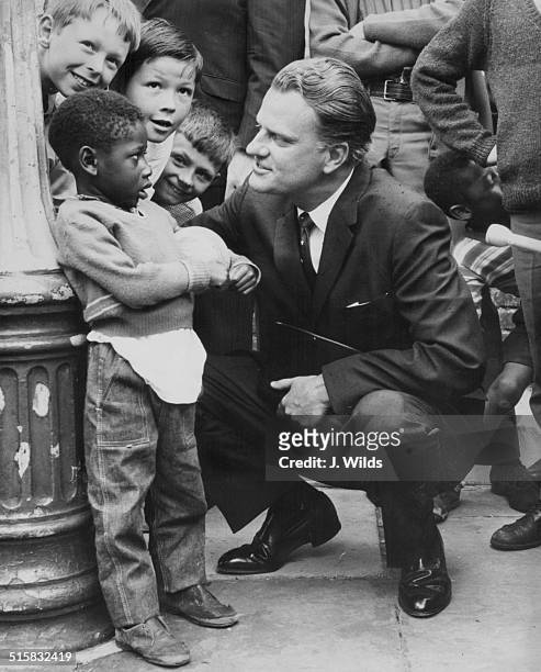American evangelist Billy Graham talking to a group of children during a visit to Brixton Parish Church, London, June 12th 1966.