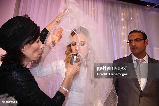 Under the Chuppa at an Orthodox Jewish wedding, the Ashkenazi bride drinks wine from a cup held by her mother as her Yemenite groom watches, Ness...