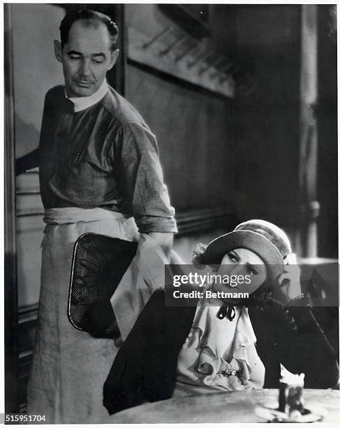 Greta Garbo seated at a table, with an actor standing behind her. Movie still from "Anna Christie." Undated photograph.