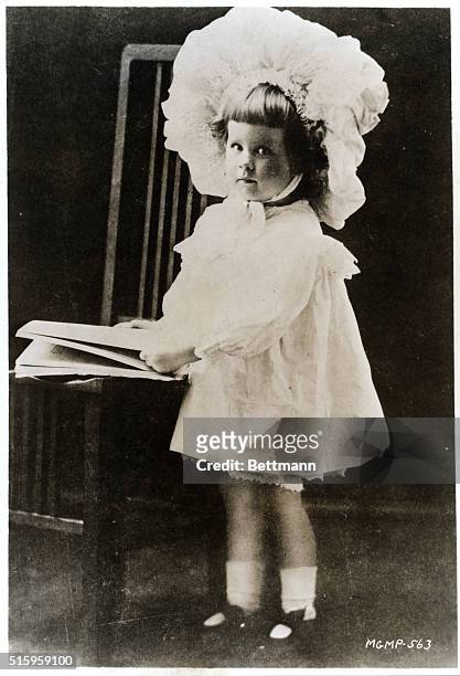 Portrait of Norma Shearer as a very young child. She wears a large baby bonnet and a long-sleeve, short, white period dress. She stands next to a...