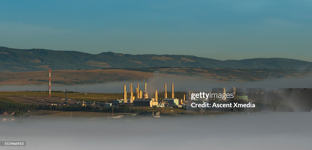 Distant gas plant emerges from fog, prairies