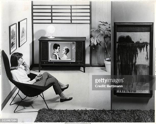 Woman watches a TV program that features a couple in a romantic mood. She sits in a reclining chair in her living room, holding the remote control....