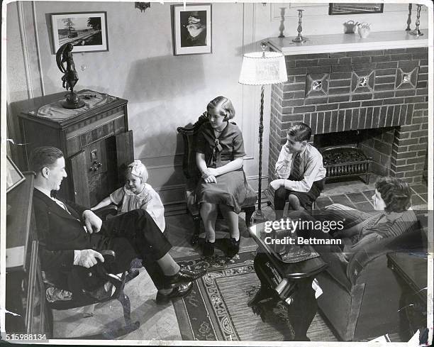 The nuclear family spends time together in their living room. The father and mother, seated in plush chairs, watch their two daughters and one son.