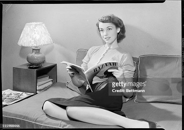 Portrait of a young woman reading Harper's Bazaar. She is shown full-length, seated on a sofa. Photo circa 1950's.