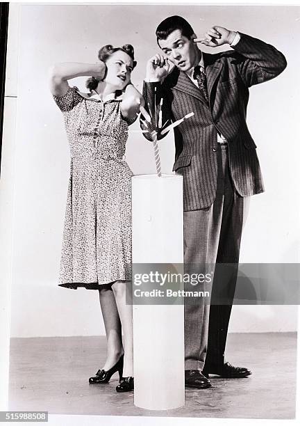 Man and woman pose with a giant firecracker. They are shown full-length, holding their ears. Undated photograph.