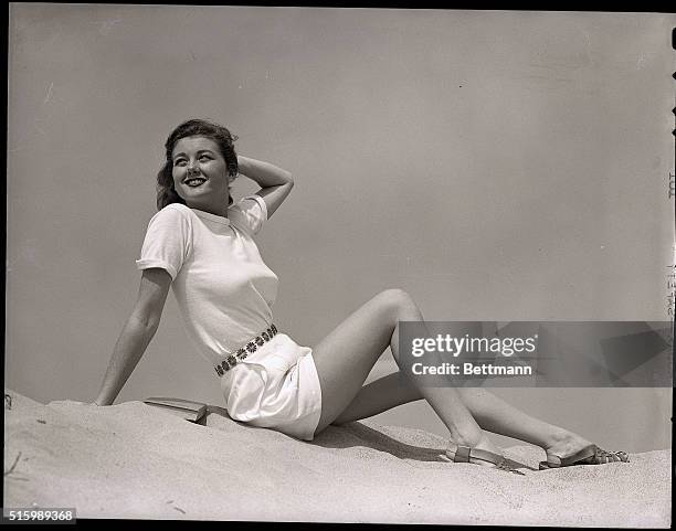 Woman on the beach, in cheesecake pose, wearing white tee-shirt and shorts with embroidered flower waistband and sandals. Ca. 1940s.