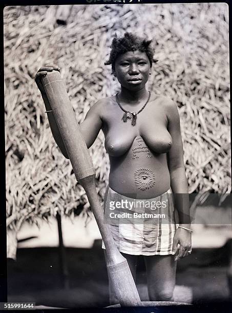 Dutch Guyanan woman is shown with ritual scarring on her shirtless chest. Her hand rests on what appears to be a large wooden tool. Undated.