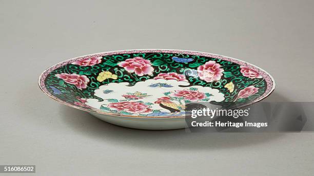 European copy of a famille noire plate with pink peonies amid floral sprays on a black glazed ground round cavetto and rim. In the centre is a petal...