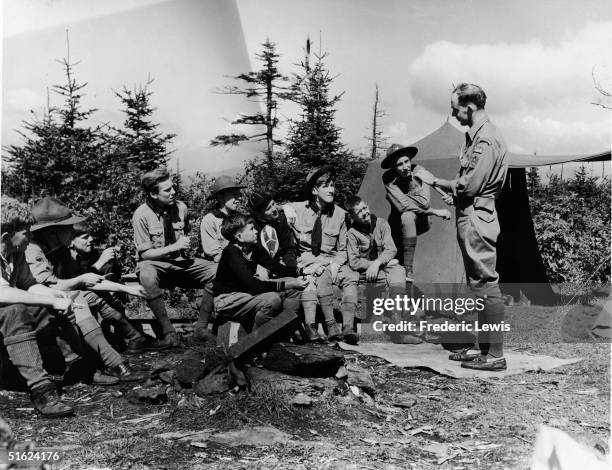Group of American boy scouts and cub scouts in uniforms that include high thick socks and various hats sit and whittle wood as they listen to their...