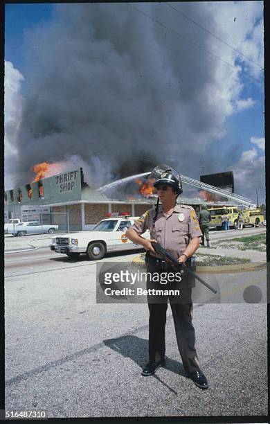 Miami: With a Dade County policeman standing guard, firemen battle a blaze at a thrift shop during the third day of racial violence. The fireman's...