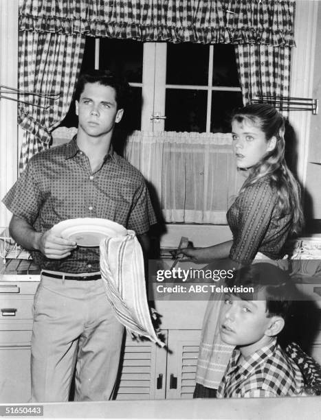 American actor Tony Dow washes dishes with a towel as he stands in the kitchen alongside an unidentified actress and Jerry Mathers who sits and gives...