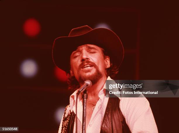 American country and western singer and guitarist Waylon Jennings wears a cowboy hat and leather vest as he sings with his eyes closed in front of a...