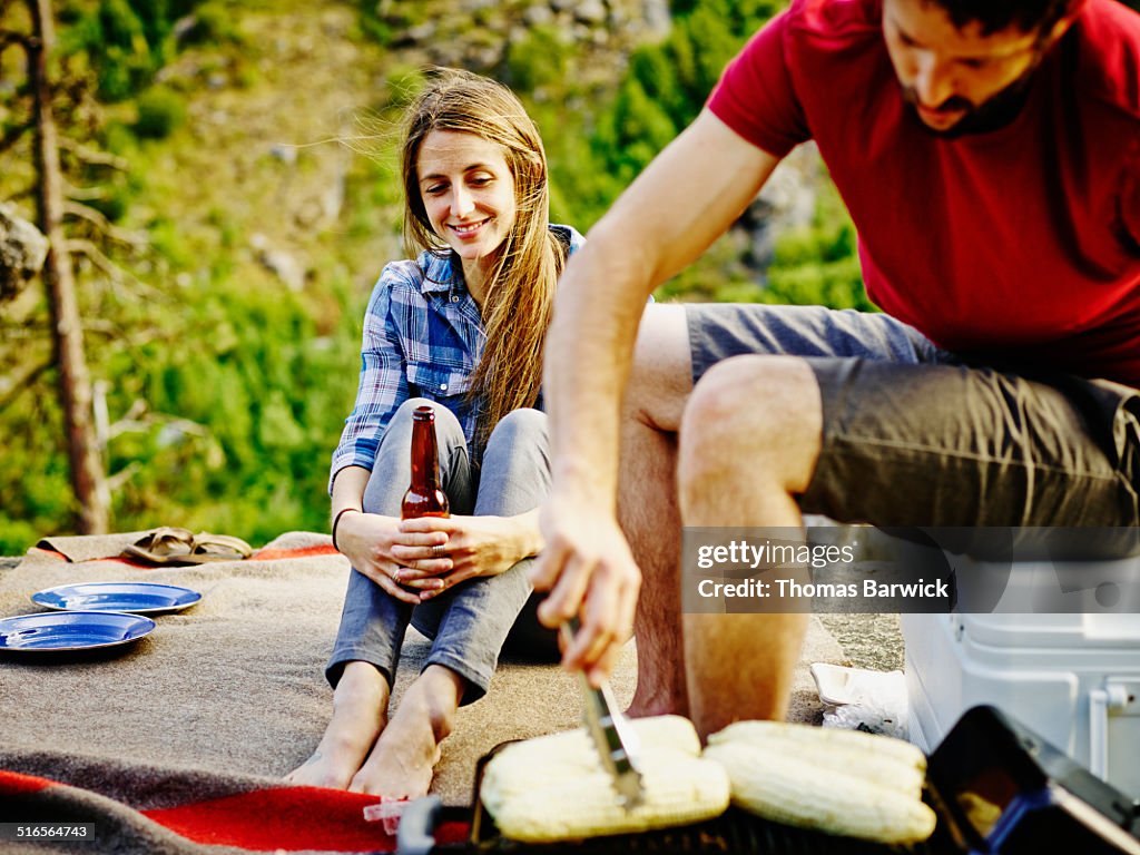 Woman sitting on blanket while boyfriend grills