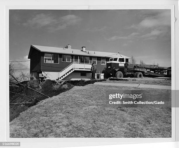 Photograph showing a landslide and ground fissure that were the result of the 1964 Alaska earthquake, a house situated at the very edge of the...