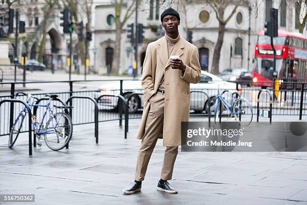 Model Adonis Bosso drinks a coffee and wears a thrift/vintage total-camel look with a peacoat, turtlneck, khaki trousers, and black slip on sneakers...