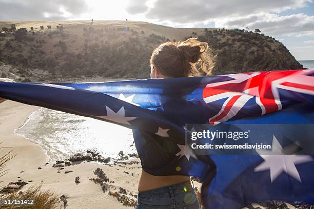 girl stands on cliff above beach holding australian's flag - australia day stock pictures, royalty-free photos & images