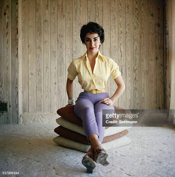 Undated photograph of actress Elizabeth Taylor, sitting on a stack of pillows, wearing purple pants and a yellow shirt, and smiling.