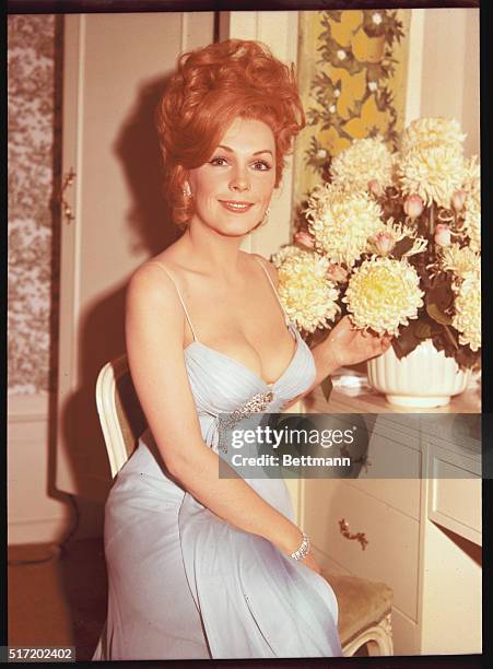 American actress Stella Stevens, born in 1936, in Mississippi. She is seated, wearing a light blue low-cut dress, pointing to a vase full of large...