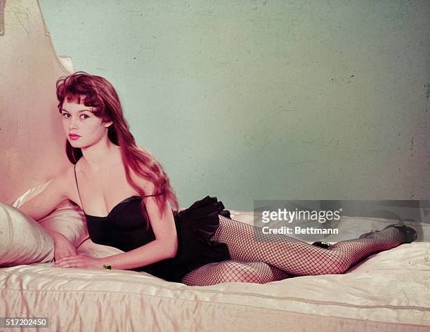 Full-length portrait of actress and sex symbol Brigitte Bardot, reclining on bed wearing a black teddy with fishnet stockings.
