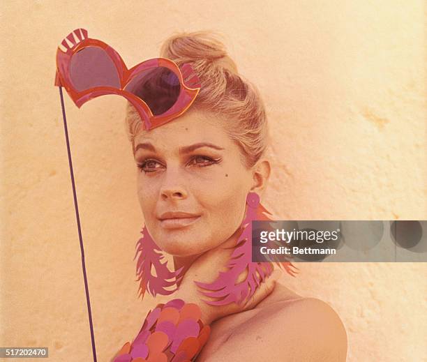 Head and shoulders photo of actress/model Candice Bergen holding a masquerade ball mask with matching, over-sized pastel earrings and bracelets.