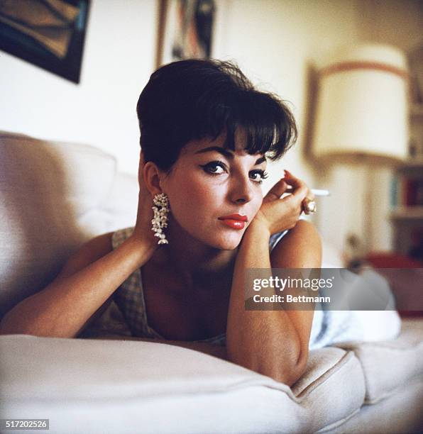 Publicity handout photograph of actress Joan Collins , on a couch, holding a cigarette near her face, ca. 1950s.