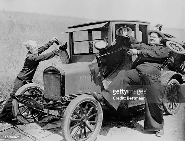 Stan Laurel and Oliver Hardy as sailors, puling the fenders off a Model T Ford while worried driver looks on. Movie still, 1928.