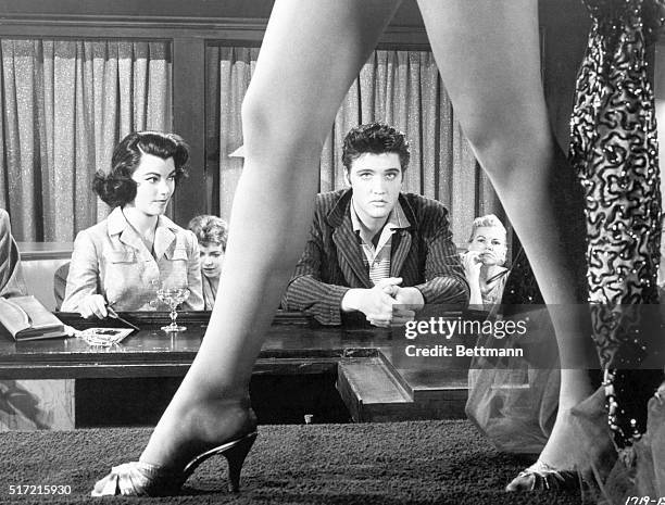 Movie still from the film "Jailhouse Rock," with Elvis Presley framed within the span of a burlesque dancer's legs as she strides the stage. Judy...