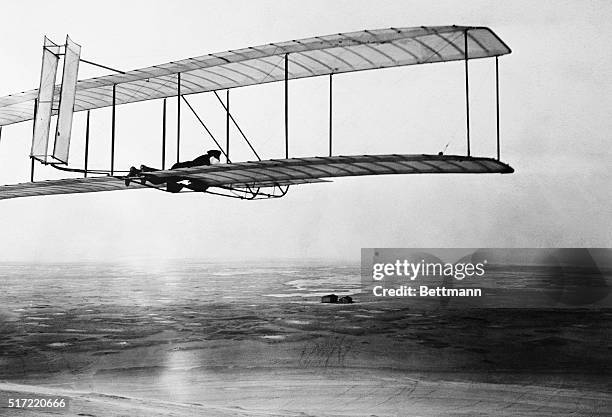 Wilbur Wright pilots a glider with a movable double rear vertical rudder over the Kill Devil Hills near Kitty Hawk. The Wright Brothers' camp can be...