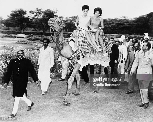 Bashir Ahmed is giving instructions to Princess Lee Radziwill and Mrs. John F. Kennedy, seated on Bashir's camel, as he prepares to lead them around...