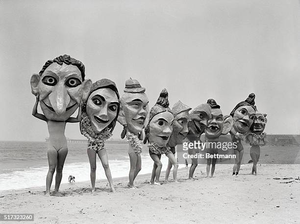 Women lined up on a beach hold giant masks in front of their faces which will be featured in the annual Venice Mardi Gras celebration.