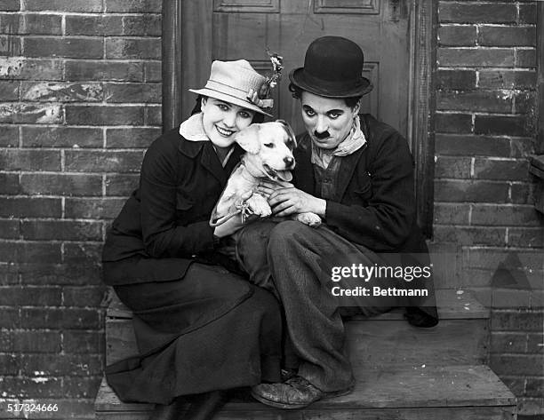 Actor Charlie Chaplin wearing his famous costume sits with actress Edna Purviance and a dog.