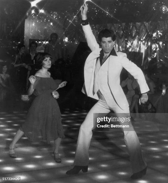 John Travolta as Tony Manero in his famous disco-dancing pose in Saturday Night Fever . His dance partner is actress Karen Lynn Gorney.