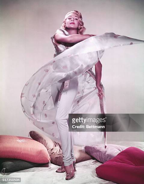 Full length portrait of American actress Lana Turner , the "Sweater Girl," wearing a white cape and standing near a pile of pillows. Undated...