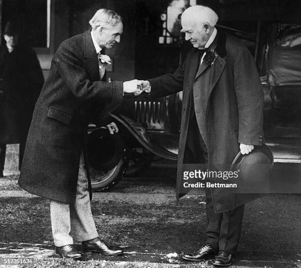 Portrait of Thomas Edison shaking hands with Henry Ford. Both are wearing overoats and flowers on their lapels. An automobile is visible in the...